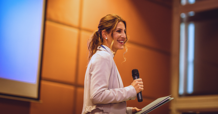 Keynote speaker smiling on stage with a microphone and notes