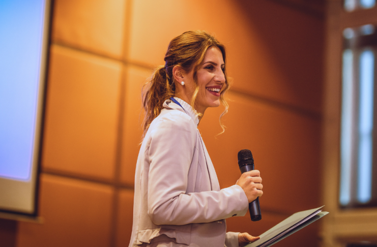 Keynote speaker smiling on stage with a microphone and notes