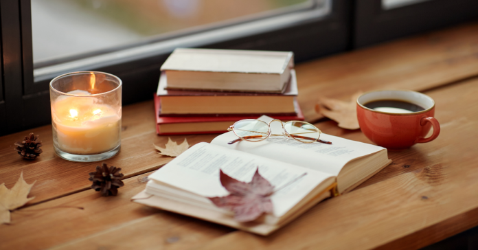 A book surrounded by fall leaves, coffee, and a candle with a view to the outdoors